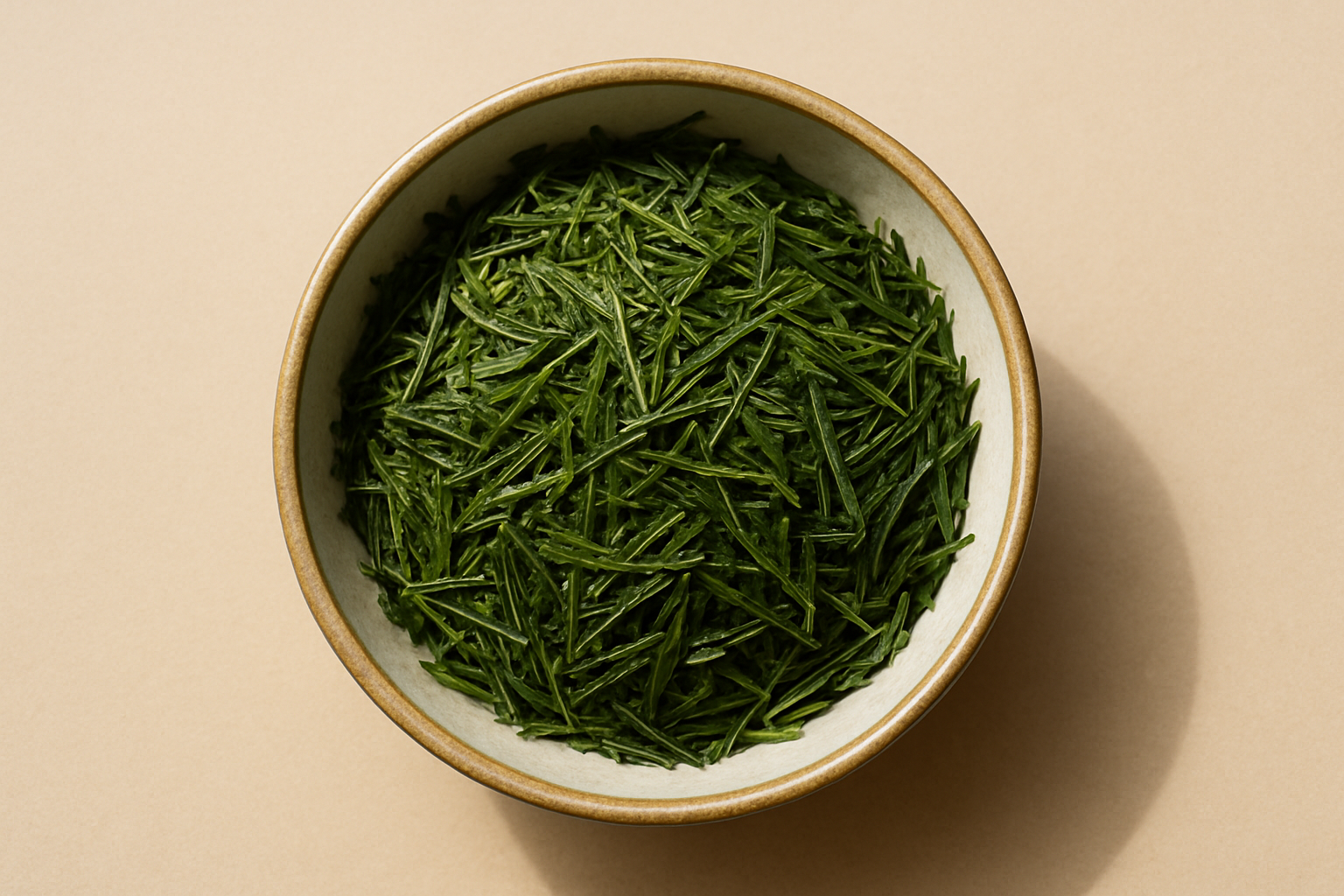 Photorealistic top-down photograph of a round beige ceramic bowl with an off-white interior, filled with loose Sencha green tea leaves (long, thin, needle-like bright green leaves). The bowl is centered on a soft matte beige background, with soft natural daylight and gentle shadows, ultra-detailed and realistic.
